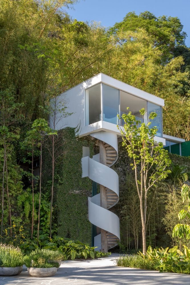 Modern white house cantilevered over a green hillside, with large glass windows and an exterior spiral staircase surrounded by vegetation.