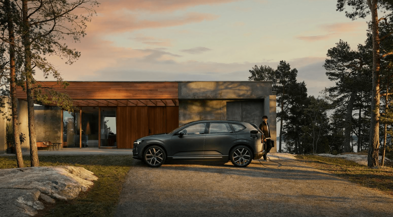 Volvo electric SUV parked in front of a modern house surrounded by trees, while a person plugs the vehicle into a charger at sunset.
