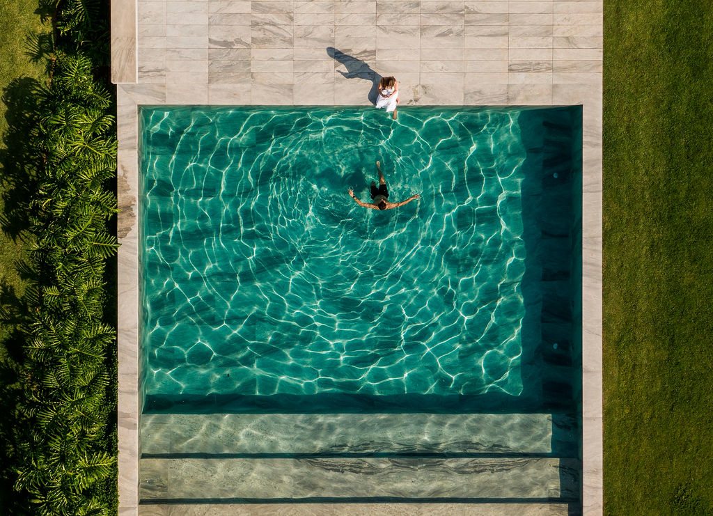 Overhead view of the swimming pool at Casa CLL by Bernardes Arquitetura, framed by white stone paving and lawn, with two people in the clear turquoise water