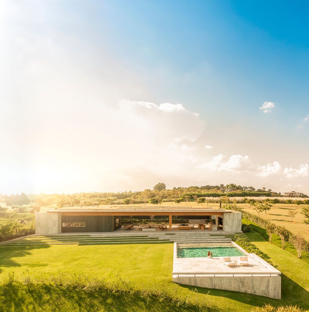 Wide view of Casa CLL by Bernardes Arquitetura at golden hour, with a green roof, open social area and swimming pool set into the sloping countryside of Porto Feliz