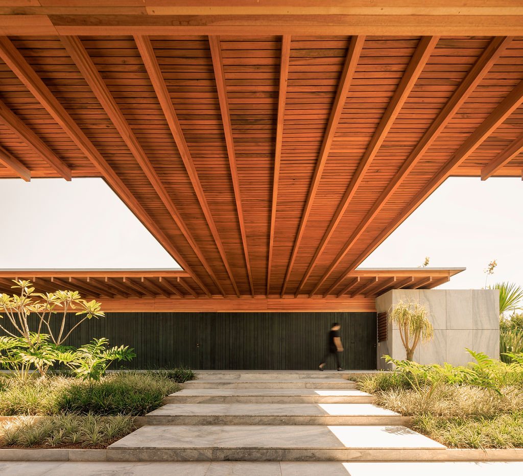 Entrance to Casa CLL by Bernardes Arquitetura, with a deep timber roof overhang, charred timber wall and stone path crossing through planted beds