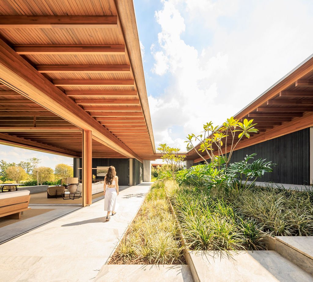Covered walkway at Casa CLL by Bernardes Arquitetura, with a timber roof on one side, open social area and a planted garden bed leading toward the landscape