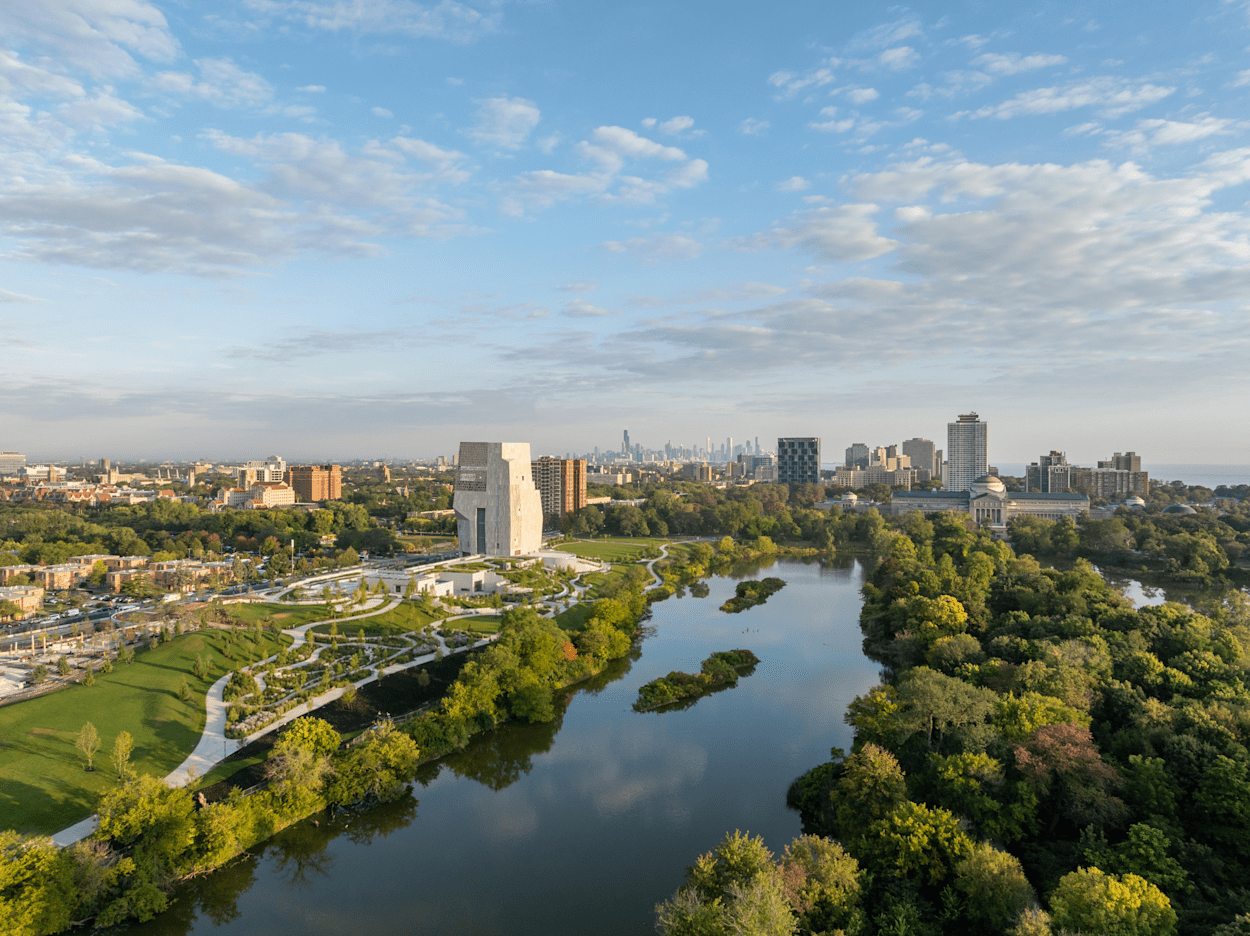 Aerial view of the Obama Presidential Center campus along Jackson Park in Chicago, featuring landscaped green spaces, a reflective lagoon, and the city skyline in the distance.