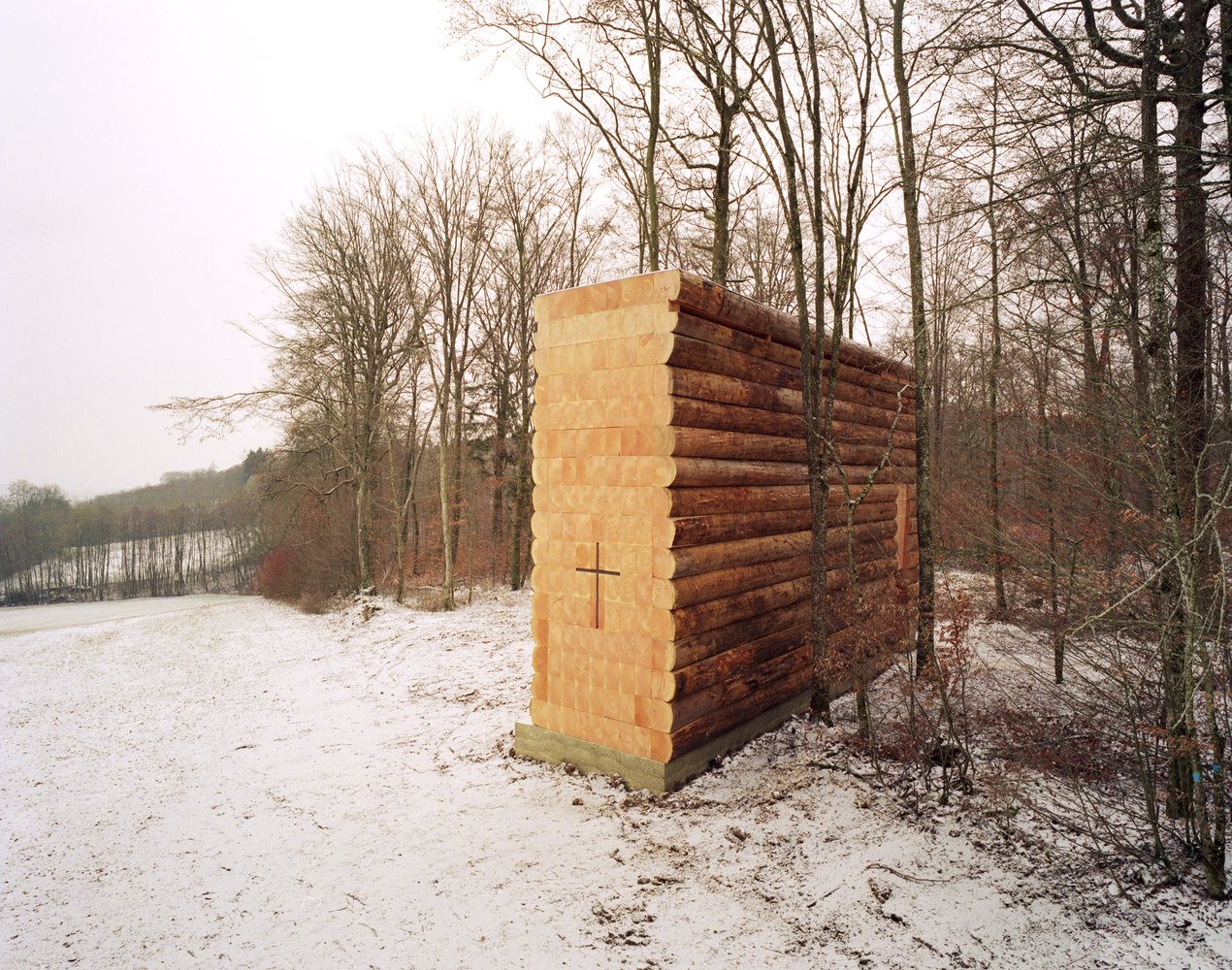 Wooden Chapel by John Pawson in snow near Unterliezheim, Germany, its stacked-timber walls and cross set against bare trees and a winter field