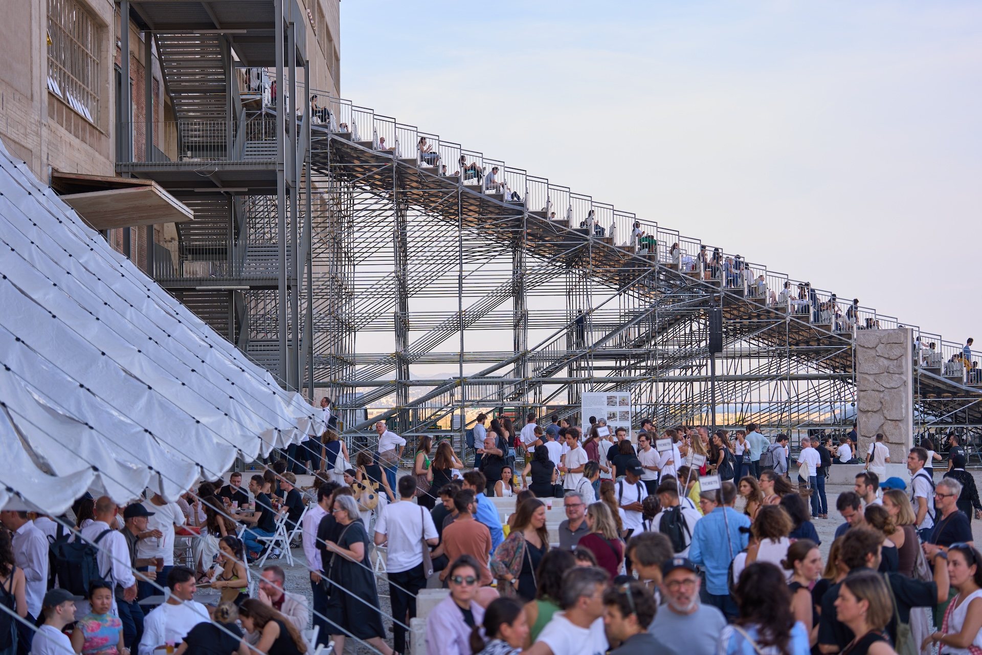 pen Forum at Tres Chimeneas during UIA Barcelona 2026, with a large scaffolding stair structure and crowds gathered outdoors at golden hour