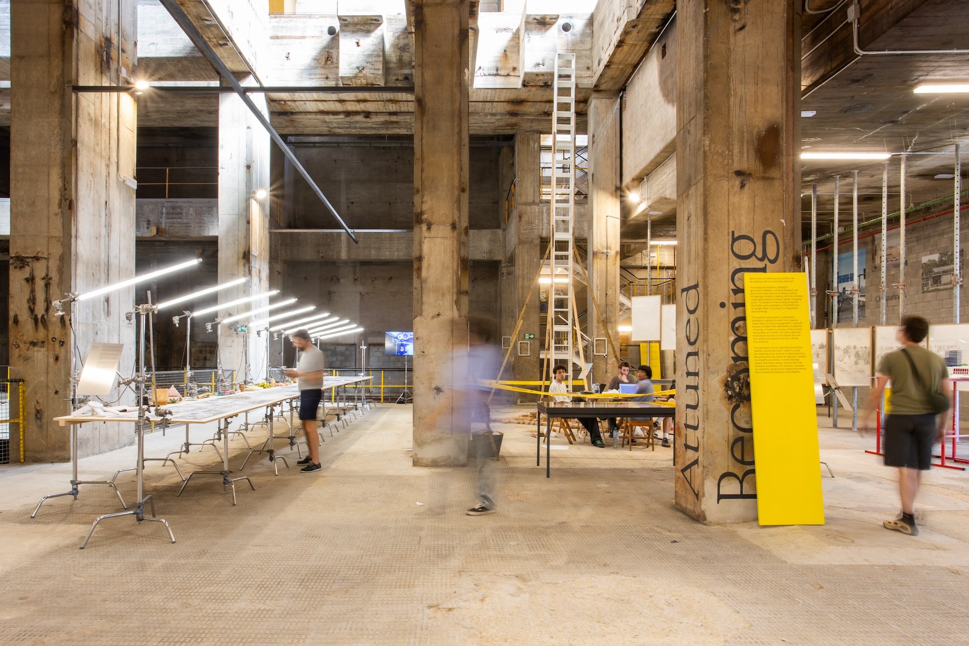 Exhibition of UIA Barcelona 2026 inside a raw concrete industrial space, with work tables, lighting rigs and yellow signage reading Attuned and Becoming