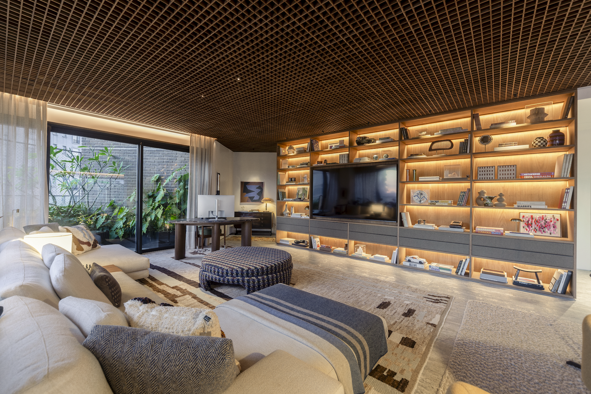 TV room at CRR | Arborea by Triplex Arquitetura, with an illuminated wooden bookshelf, sofa, workspace and a green patio view in Itaim Bibi, São Paulo