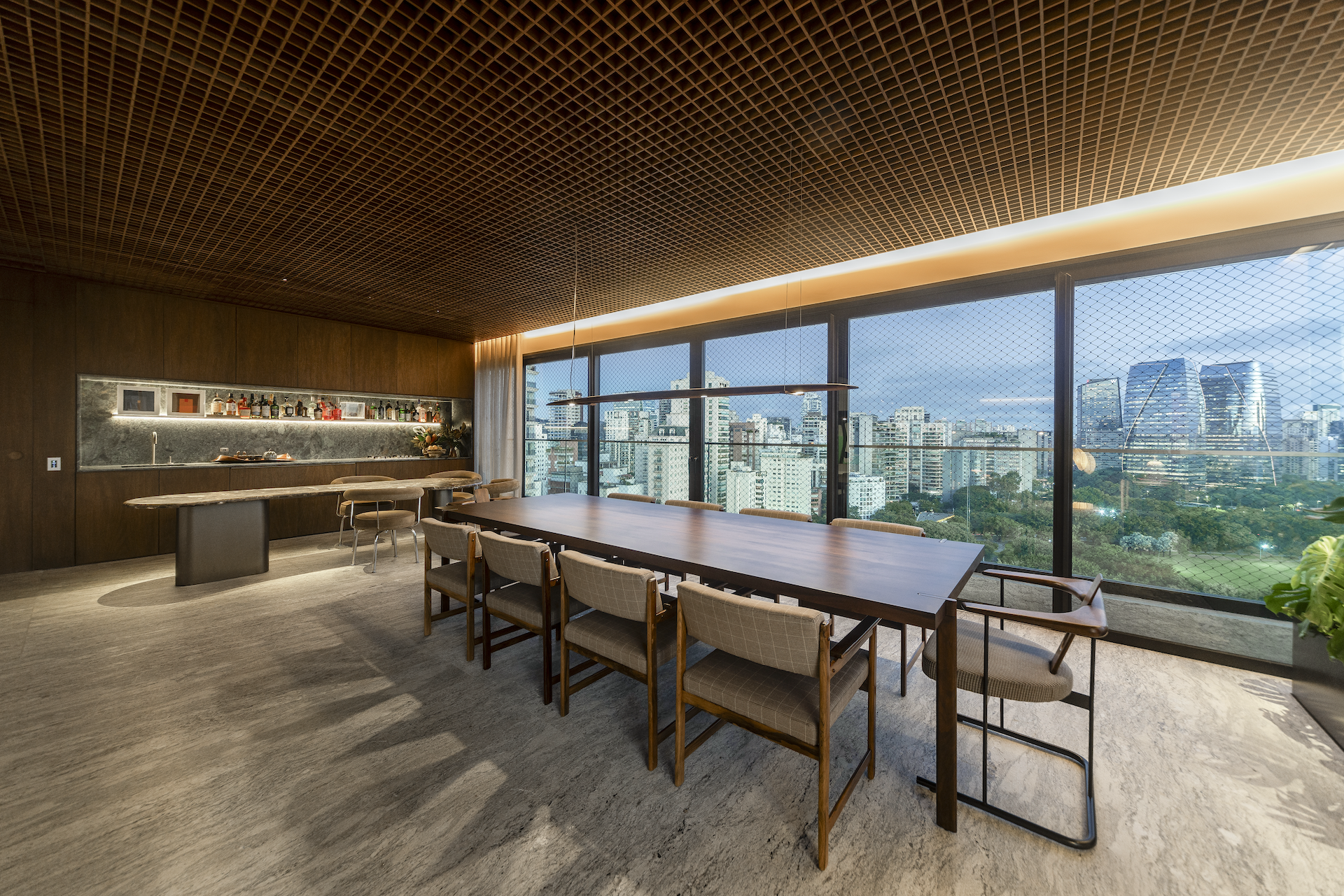 Dining and gourmet area at CRR | Arborea by Triplex Arquitetura at dusk, with a long wooden table, upholstered chairs, stone bar and São Paulo skyline in Itaim Bibi