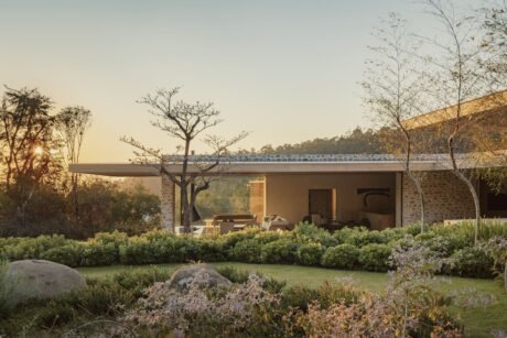 Casa Grama by Studio Arthur Casas in Itupeva, São Paulo, low stone-and-glass volume open to the garden at sunset, with clipped hedges, boulders and slender trees in the foreground
